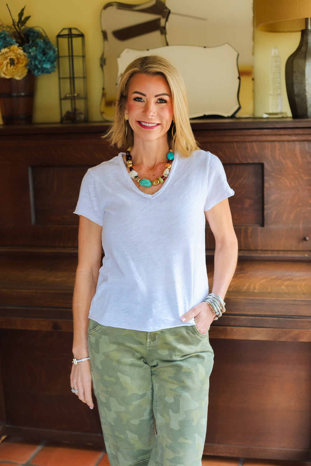 Woman wearing a light blue shirt and green camouflage pants standing in a room with wooden furniture and decorative items.
