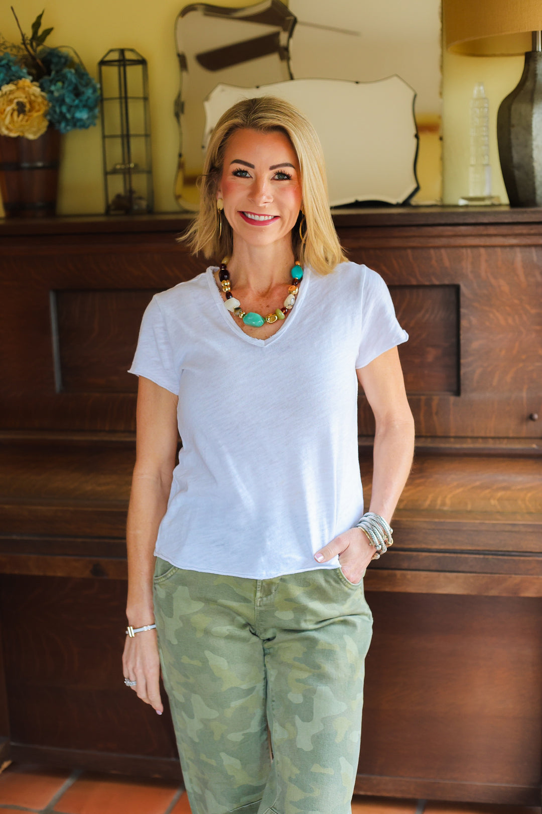 Woman wearing a light blue shirt and green camouflage pants standing in a room with wooden furniture and decorative items.
